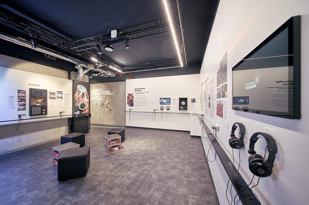 Innenansicht der Blindenfußball-Ausstellung im FC St. Pauli-Museum mit Ausstellungswänden, Bildschirmen, Hörstationen und Sitzgelegenheiten.