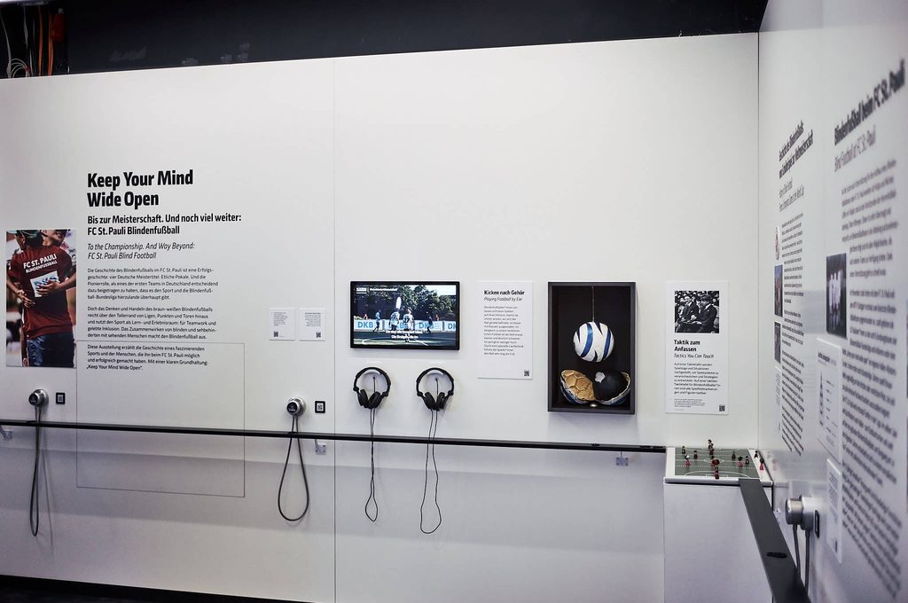 Ausstellungswand „Keep Your Mind Wide Open“ über den Blindenfußball beim FC St. Pauli mit Bildschirm, Kopfhörern, Fußbällen und Informationstafeln.